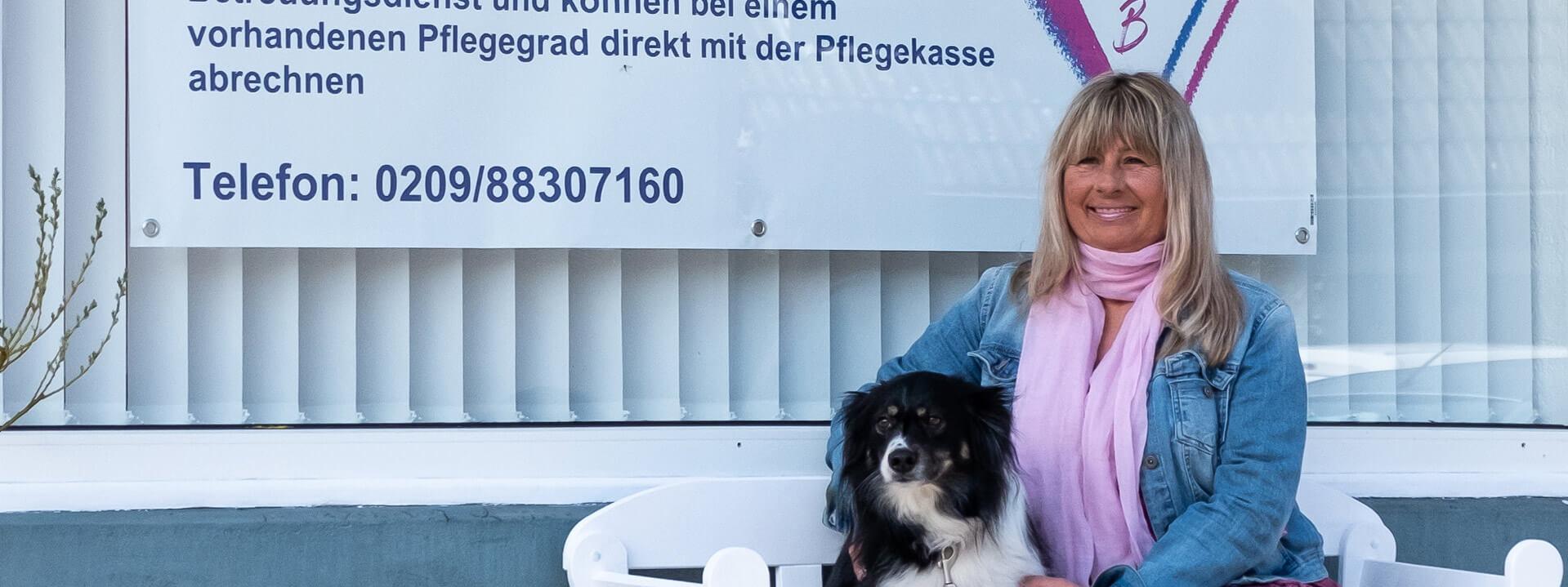 Heike Becker, eine Frau mittleren Alters, sitzt mit einem Hund vor einem Schild mit der Aufschrift "Seniorenbetreuung mit Herz".