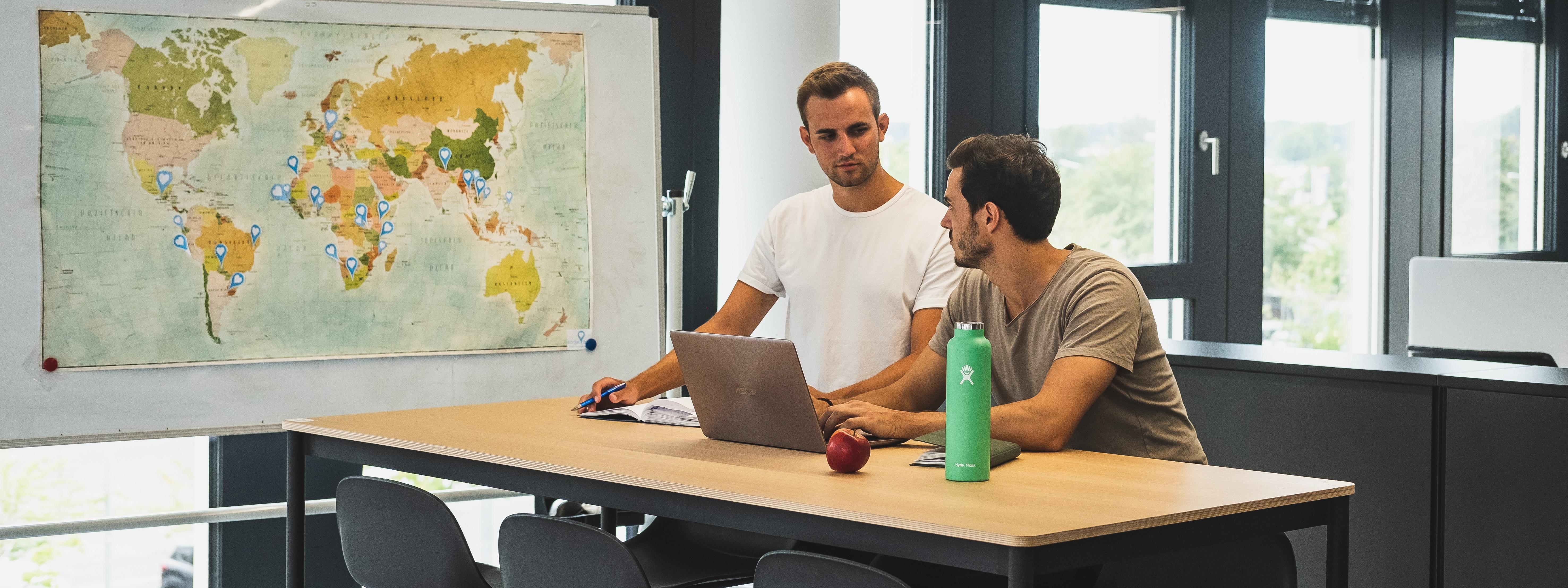 Die Gründer von Socialbnb stehen an einem Schreibtisch, neben ihnen eine Weltkarte