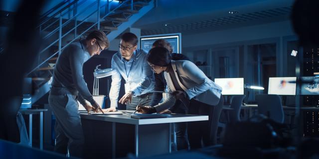 Four people standing in an office are looking on a laptop screen