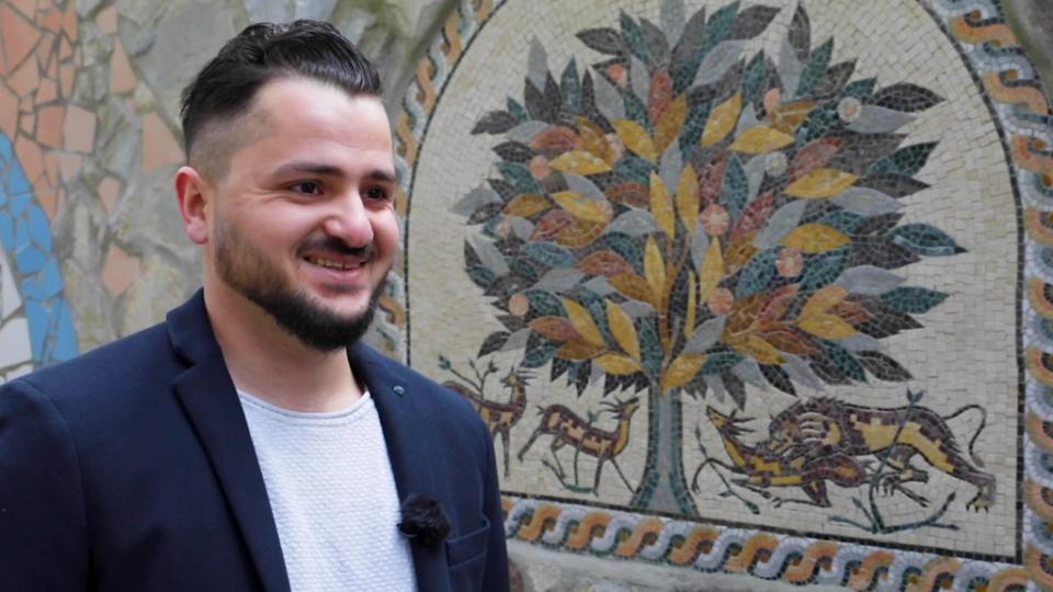 Houran Almahrouk, ein junger Mann mit Bart, steht vor einem Wandmosaik mit einem Baummotiv.