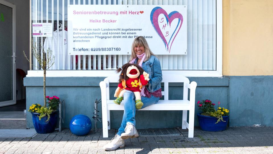 Heike Becker, eine Frau mittleren Alters, sitzt mit einer Handpuppe auf einer Bank vor einem Schild mit der Aufschrift "Seniorenbetreuung mit Herz".