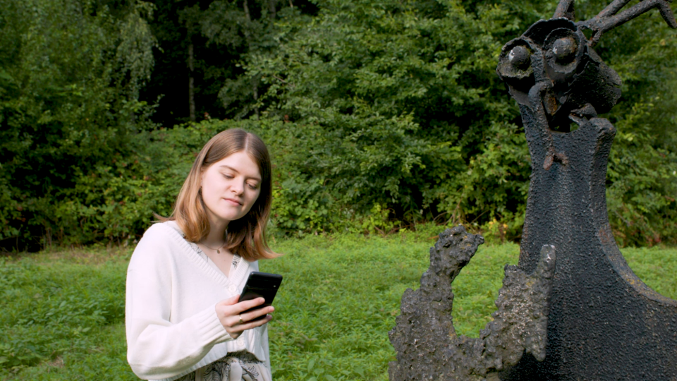 Elisabeth Jacobsohn is standing in a park next to a scalpture looking on her smartphone