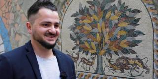Houran Almahrouk, ein junger Mann mit Bart, steht vor einem Wandmosaik mit einem Baummotiv.