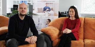 A man with a bald head and a full beard and a woman sitting together on sofas.