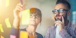 Frau und Mann stehen vor einem Glasboard, auf dem bunte Post-its kleben