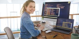 Frau sitzt in einem Büro am Schreibtisch vor einem Laptop und einem Desktop-Bildschirm.