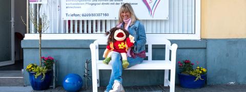 Heike Becker, eine Frau mittleren Alters, sitzt mit einer Handpuppe auf einer Bank vor einem Schild mit der Aufschrift "Seniorenbetreuung mit Herz".