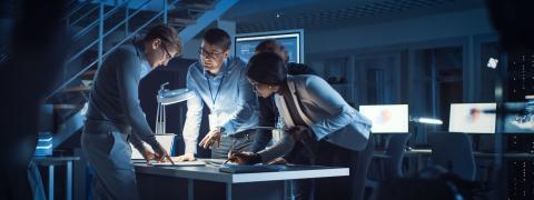 Four people standing in an office are looking on a laptop screen