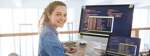 Frau sitzt in einem Büro am Schreibtisch vor einem Laptop und einem Desktop-Bildschirm.