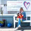 Heike Becker, eine Frau mittleren Alters, sitzt mit einer Handpuppe auf einer Bank vor einem Schild mit der Aufschrift "Seniorenbetreuung mit Herz".