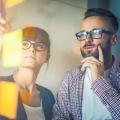 Frau und Mann stehen vor einem Glasboard, auf dem bunte Post-its kleben