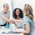 Drei Frauen und ein Mann am Konferenztisch. Zwei Frauen geben sich die Hand.