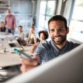 Mann steht am Whiteboard und schreibt etwas darauf. Dahinter eine Gruppe Personen am Konferenztisch