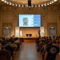 Großer Saal mit Publikum. Ein Mann hält einen Vortrag vor einem großen Bildschirm, der an der Wand hängt.